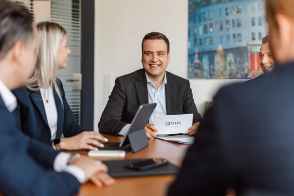 Fünf Personen in Businesskleidung sitzen an einem Konferenztisch, ein Mann hält ein Blatt mit dem Logo 'DHS' und lächelt