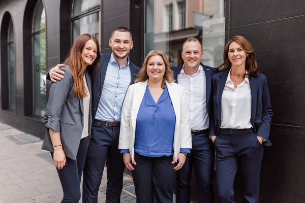 Fünf beruflich gekleidete Personen stehen eng zusammen vor einem Gebäude und lächeln in die Kamera