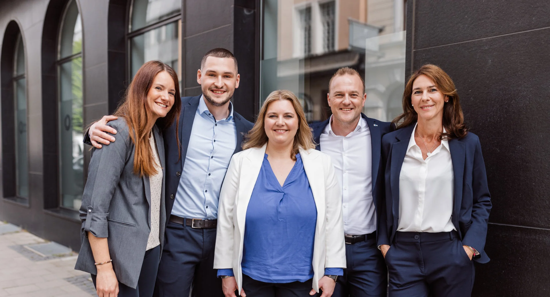 Fünf beruflich gekleidete Personen stehen eng zusammen vor einem Gebäude und lächeln in die Kamera