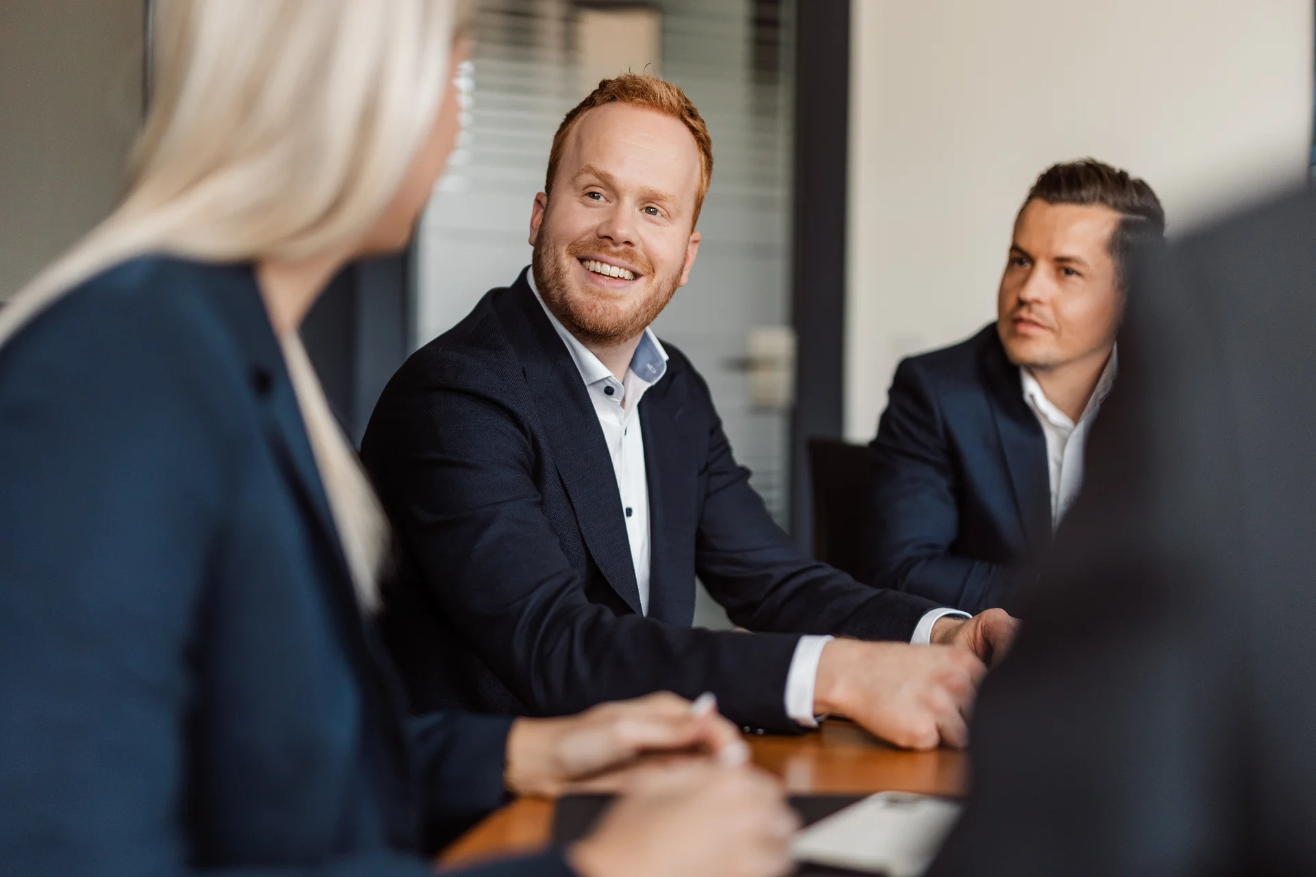 Es sind drei Personen in einer Besprechung und in Businesskleidung zu sehen, der Hinterkopf einer Frau, ein Mann der sie anlächelt und ein Mann der seitloch sitzt und ebenfalls lächelt